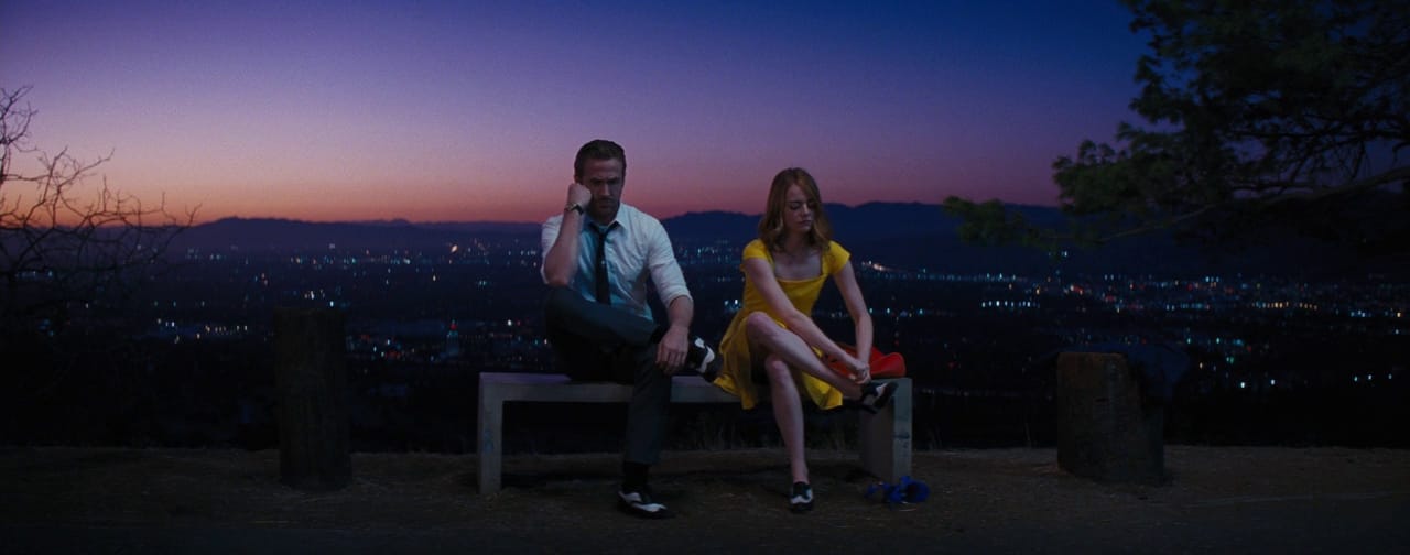 A man and woman sit on a bench with a backdrop of the city during sunset.