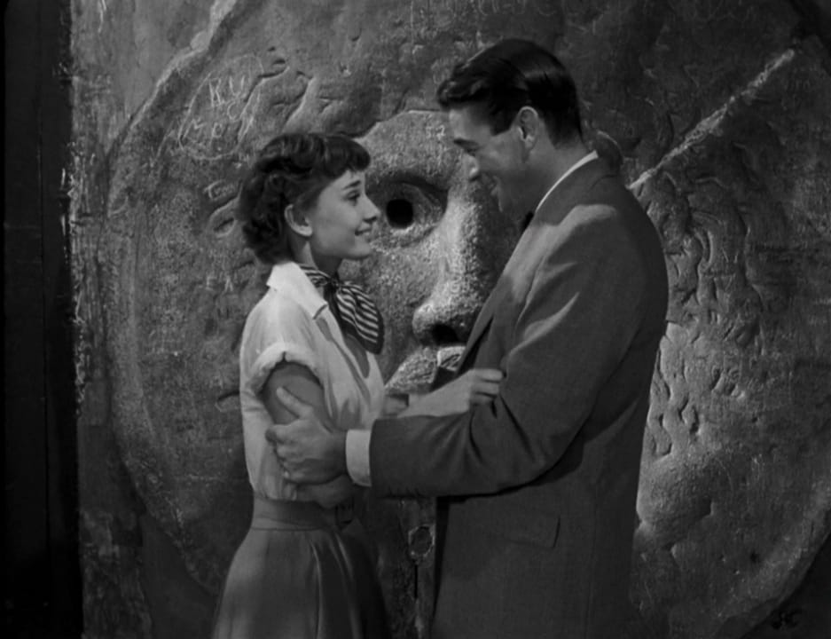 A man and a woman hold each other in front of a statue of a face.